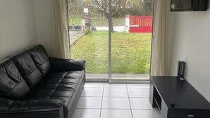 A modern living room with a black leather sofa, a small black table, and a sliding glass door leading to a garden. - Our gîtes are ideally located in Pontonx-sur-l'Adour (Pontonx-Sur-L'adour)