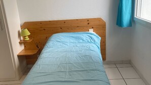 A small, single-bed room with a wooden headboard, a bedside table with a lamp, and a window with a blue curtain. - Our gîtes are ideally located in Pontonx-sur-l'Adour (Pontonx-Sur-L'adour)