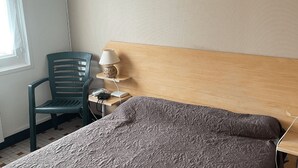 Een netjes ingerichte hotelkamer met een enkel bed, een stoel, een nachtkastje met lamp en een klein plankje.