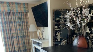 Una habitación con piso de madera, una mesa con superficie negra sobre blanca, un mueble blanco, un televisor montado en la pared y un jarrón decorativo con flores.