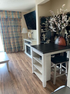 Une pièce avec un plancher en bois, une table blanche avec un plateau noir, un meuble blanc, un téléviseur fixé au mur et un vase décoratif avec des fleurs.