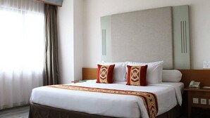 A hotel room with a large bed, two decorative pillows, a bedside table with a phone, and a window with curtains. - Asana Kawanua Jakarta (Jakarta)