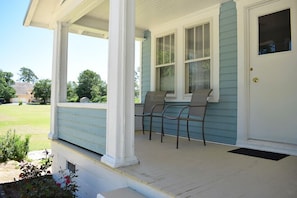Eine Veranda mit zwei Stühlen und einer Tür, ein Rasen und ein blaues Haus.