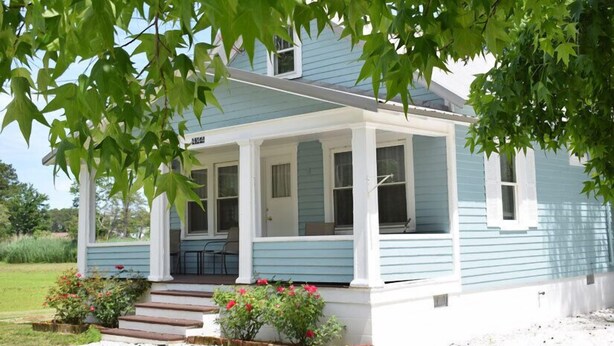 Ein hellblaues Haus mit Veranda, Stufen zum Eingang und einem Garten mit roten Blumen.