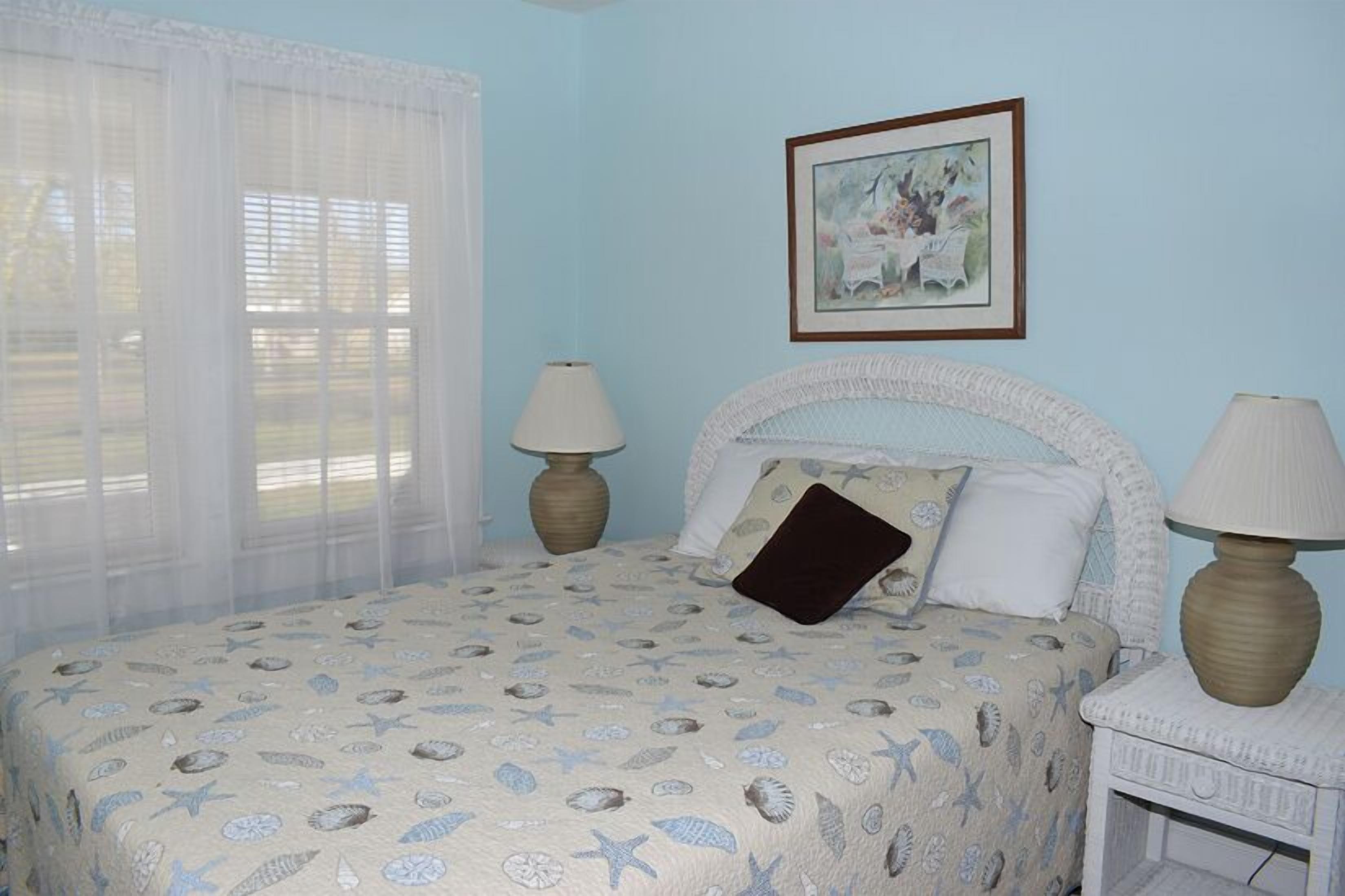 Une chambre à coucher avec un lit, deux lampes de chevet, un tableau encadré au mur et des rideaux légers à la fenêtre.