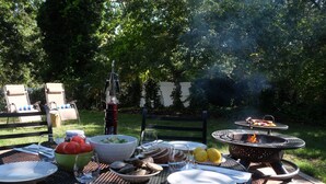 An outdoor dining area with a round table set for a meal, surrounded by greenery and a fire pit. - Charming, newly renovated house, a few steps from bike path. (Dennis Port)