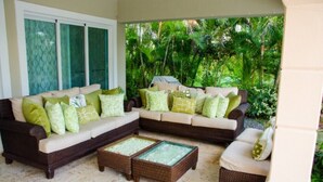 A covered outdoor seating area with a sofa and armchairs, surrounded by lush greenery. - Villa Esmeralda Cocotal Golf & Country (Punta Cana)