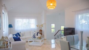 A spacious living room with a white sofa set, a glass coffee table, and a television. - Villa Esmeralda Cocotal Golf & Country (Punta Cana)