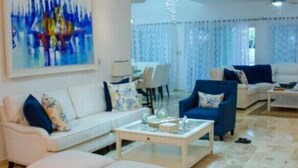 A modern living room with a white sofa, blue armchair, and ottoman, a coffee table, and a chandelier. - Villa Esmeralda Cocotal Golf & Country (Punta Cana)