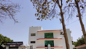 The exterior of Hotel Corbett Regency, featuring a modern design with a white facade and red accents, a gated entrance, and a parking area with a car. - Deluxe family Balcony (Ramnagar)
