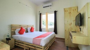 A room with a wooden bed, a small bedside table, a TV mounted on a wooden cabinet, and a window with curtains. - Deluxe family Balcony (Ramnagar)