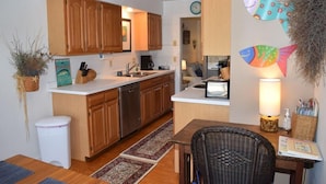 A compact kitchen with wooden cabinets, a white countertop, and a dining area with a wicker chair.