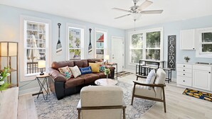 A spacious living room with a brown leather sofa, a beige armchair, and a coffee table. There is a ceiling fan, a dining area with a table and chairs, and a kitchen with white cabinets.