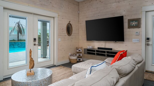 Een moderne woonkamer met een beige bank, een flatscreen televisie en uitzicht op een zwembad door glazen deuren.