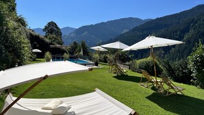 Un área de piscina con sillones de descanso y sombrillas, rodeada de exuberante vegetación y montañas.