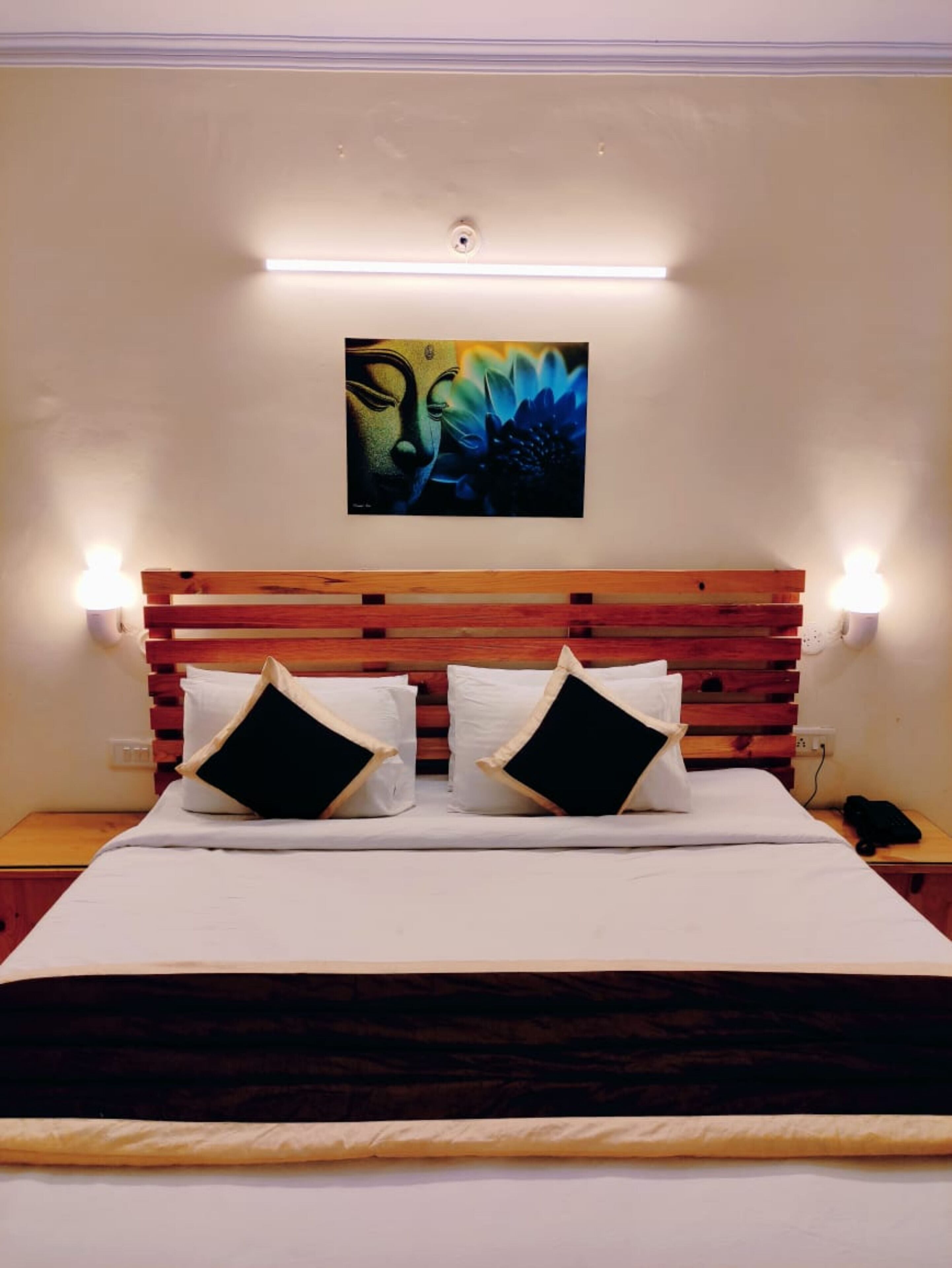 Una habitación de hotel con cabecera de madera, dos lámparas de noche, una cama con ropa de cama blanca y almohadas negras, y un cuadro en la pared.