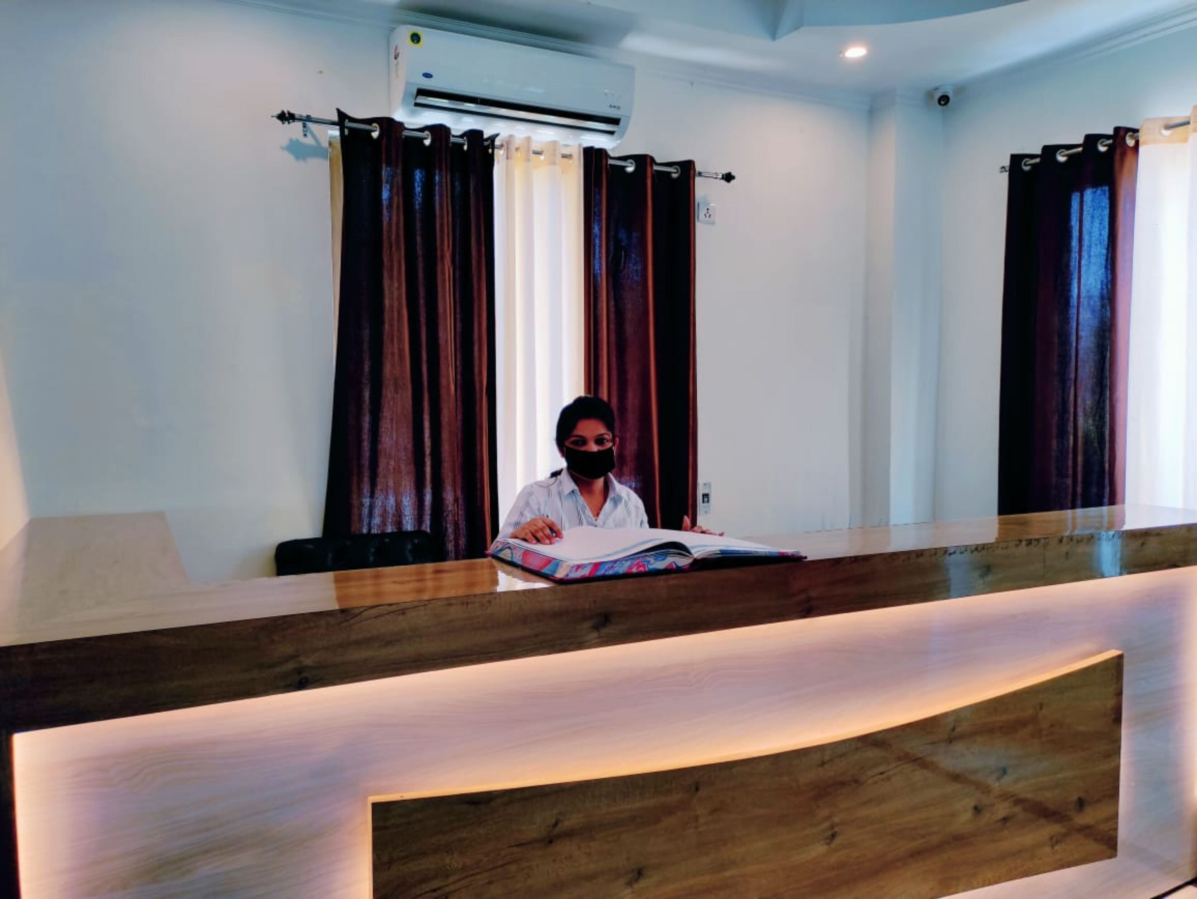 A reception desk with a wooden counter, a person seated behind it, and a window with curtains.