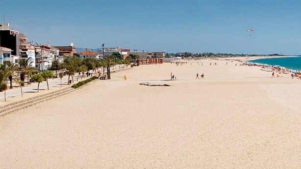 Ein weiter Sandstrand mit blauem Himmel, Palmen und Gebäuden im Hintergrund.