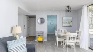Una sala de estar moderna con un sofá gris, una mesa de comedor blanca con sillas, una lámpara y un cuadro en la pared.