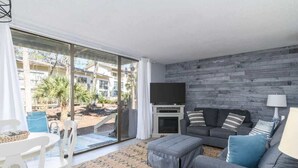 Una sala de estar moderna con un sofá gris, un televisor de pantalla plana y una chimenea. Hay un amplio ventanal corredero que da a un espacio exterior con plantas y vistas a edificios.