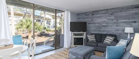 A modern living room with a grey sofa, a flat-screen TV, and a fireplace. There is a large sliding glass door leading to an outdoor area with plants and a view of buildings.