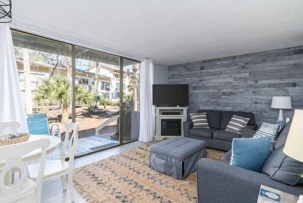 Una sala de estar moderna con un sofá gris, un televisor de pantalla plana y una chimenea. Hay un amplio ventanal corredero que da a un espacio exterior con plantas y vistas a edificios.