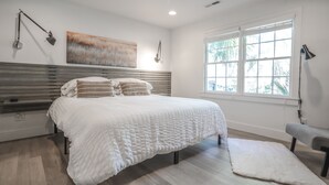 A modern bedroom with a large bed, a bench, a window with blinds, and a painting on the wall.