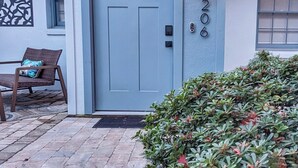 A house with a light blue door, a number '206' on the wall, a wicker chair with a cushion, and a small garden.
