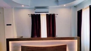 A reception desk with a wooden finish and integrated lighting, set against a white wall with a ceiling light fixture. - Family Suite With Balcony 2 (Ramnagar)