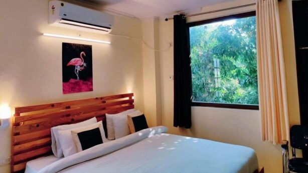 A hotel room with a wooden headboard, a bed with white linens, a wall-mounted air conditioner, a window with curtains, and a painting of a flamingo. - Family Suite With Balcony 2 (Ramnagar)