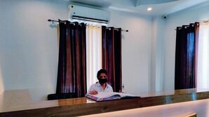 A reception desk with a person seated behind it, a wooden counter, and a wall-mounted air conditioning unit. - Family Suite With Balcony 2 (Ramnagar)