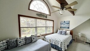 Ein Schlafzimmer mit schrägen Wänden, einem Deckenventilator, einem großen Fenster, zwei Betten mit gemusterten Bettbezügen und einem Bild an der Wand.