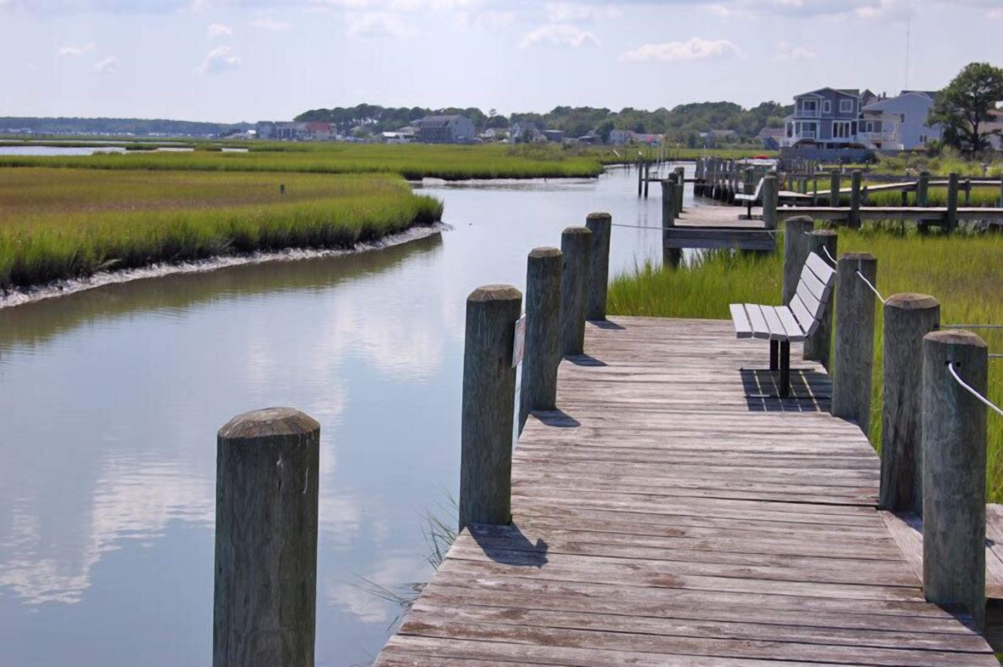 A wooden pier leading to a bench overlooking a calm river, with houses in the background.
