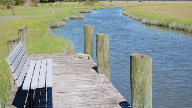 Eine Holzstege mit einer Bank erstreckt sich über einen ruhigen Fluss, umgeben von grasbewachsenen Ufern und Bäumen im Hintergrund.
