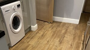 A laundry area with a washing machine, wooden flooring, and a door leading to another room. - Large 3 bed House Sleep 10 Anfield Liverpool (Liverpool)