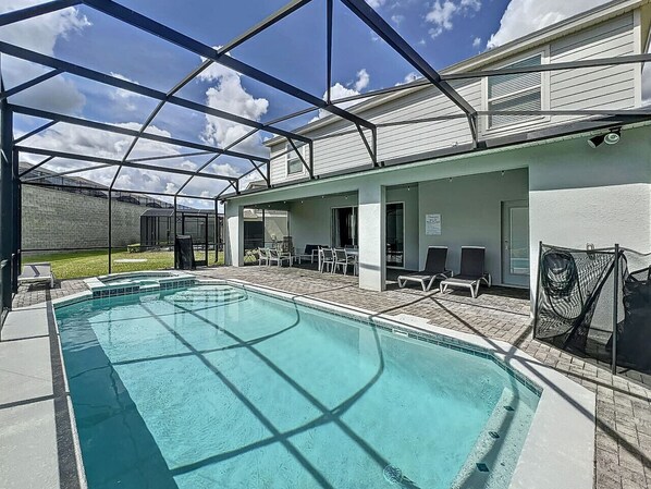 A swimming pool with a covered patio area, lounge chairs, and a view of a building with a brick wall and a door.