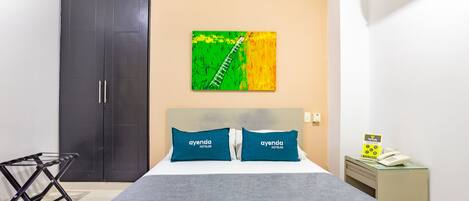 A hotel room with a bed, two pillows, a wall-mounted artwork, a wardrobe, a desk with a telephone, and a hanging laundry rack.