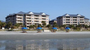 Hotel a peu de platja amb diverses plantes, balcons i ombrel·les blaves a la platja.