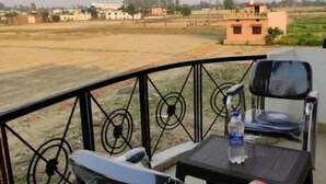 A balcony with two chairs and a table, overlooking a rural landscape with fields and buildings. - Suite with Balcony 2 (Ramnagar)