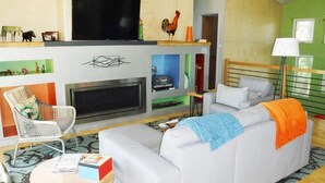 A modern living room with a sofa, a white chair, a flat-screen TV, a fireplace, and a bookshelf. - Tin Cup Palace- Community Boat Launch, Fire Pit & Screened Porch (Chincoteague)