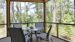 A covered outdoor seating area with a glass table and chairs, surrounded by a wooden railing, overlooking a forested area. - Tin Cup Palace- Community Boat Launch, Fire Pit & Screened Porch (Chincoteague)
