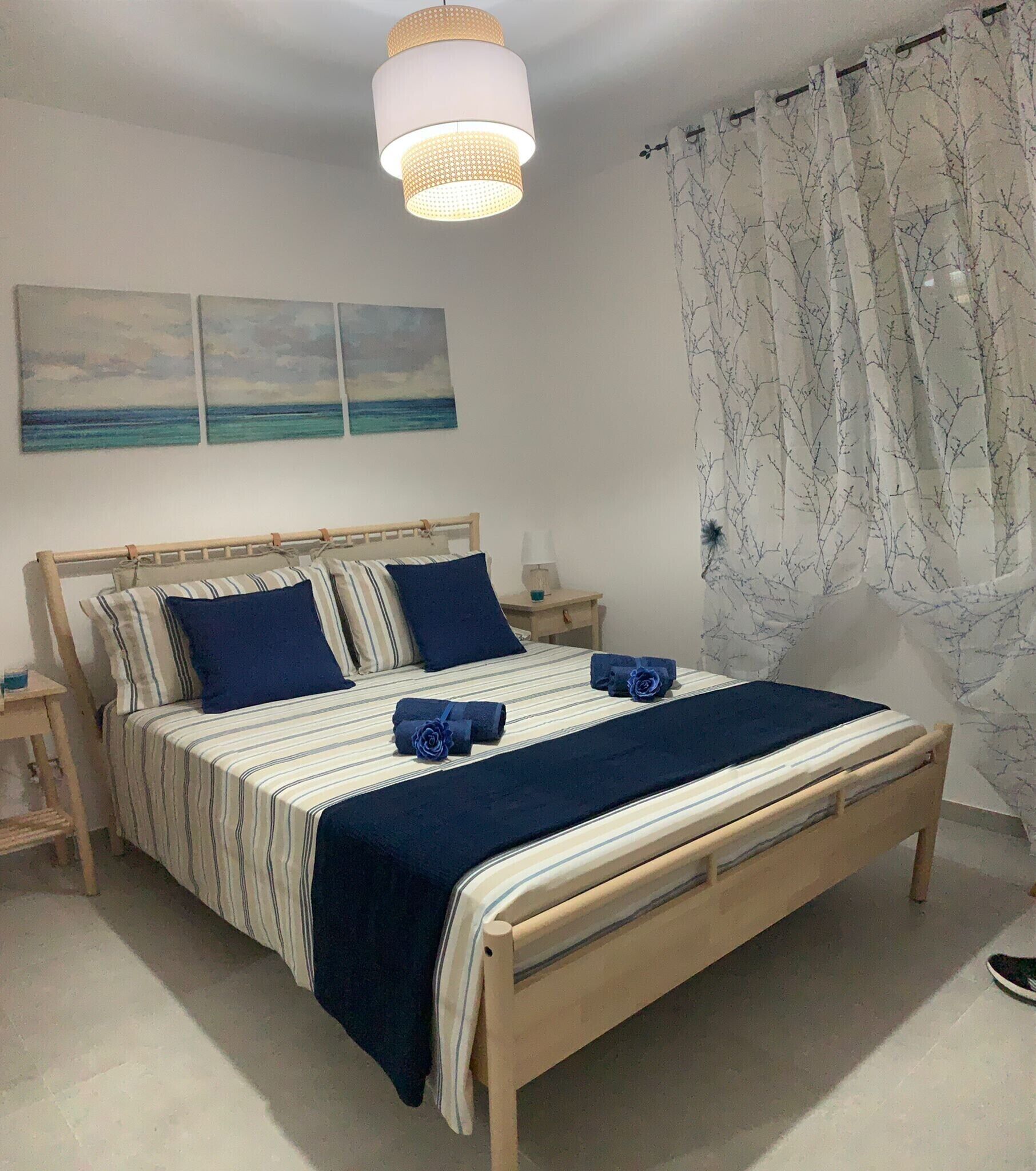 Een net opgemaakt bed met een gestreept dekbed en blauwe kussens, een nachtkastje met een lamp, en uitzicht op zee vanuit het raam.