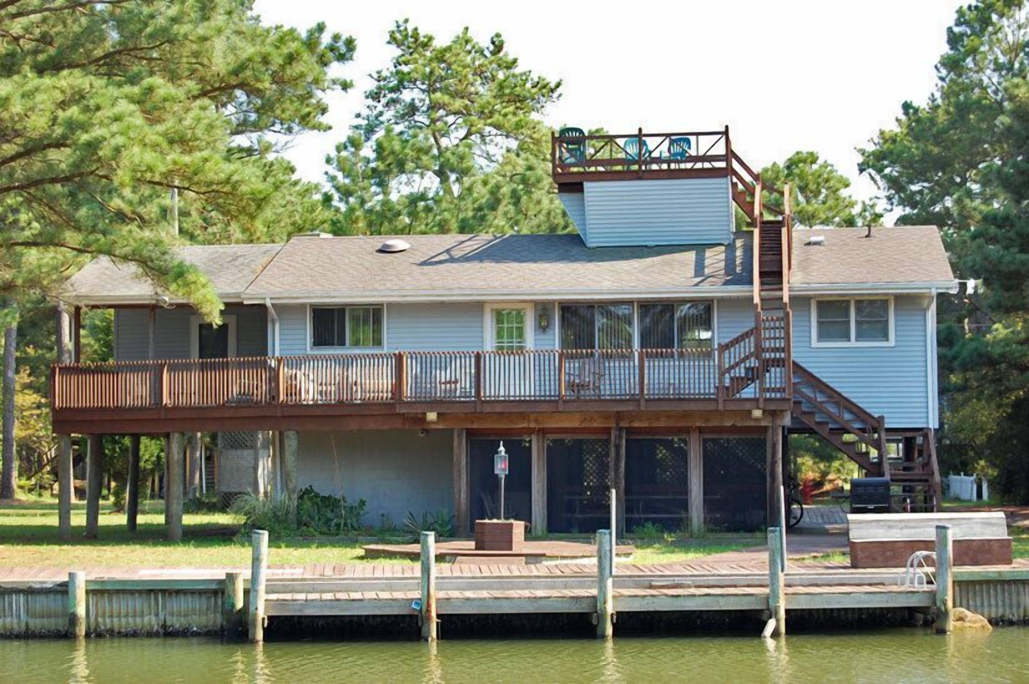 A two-story house with a dock extending into a body of water, surrounded by trees.