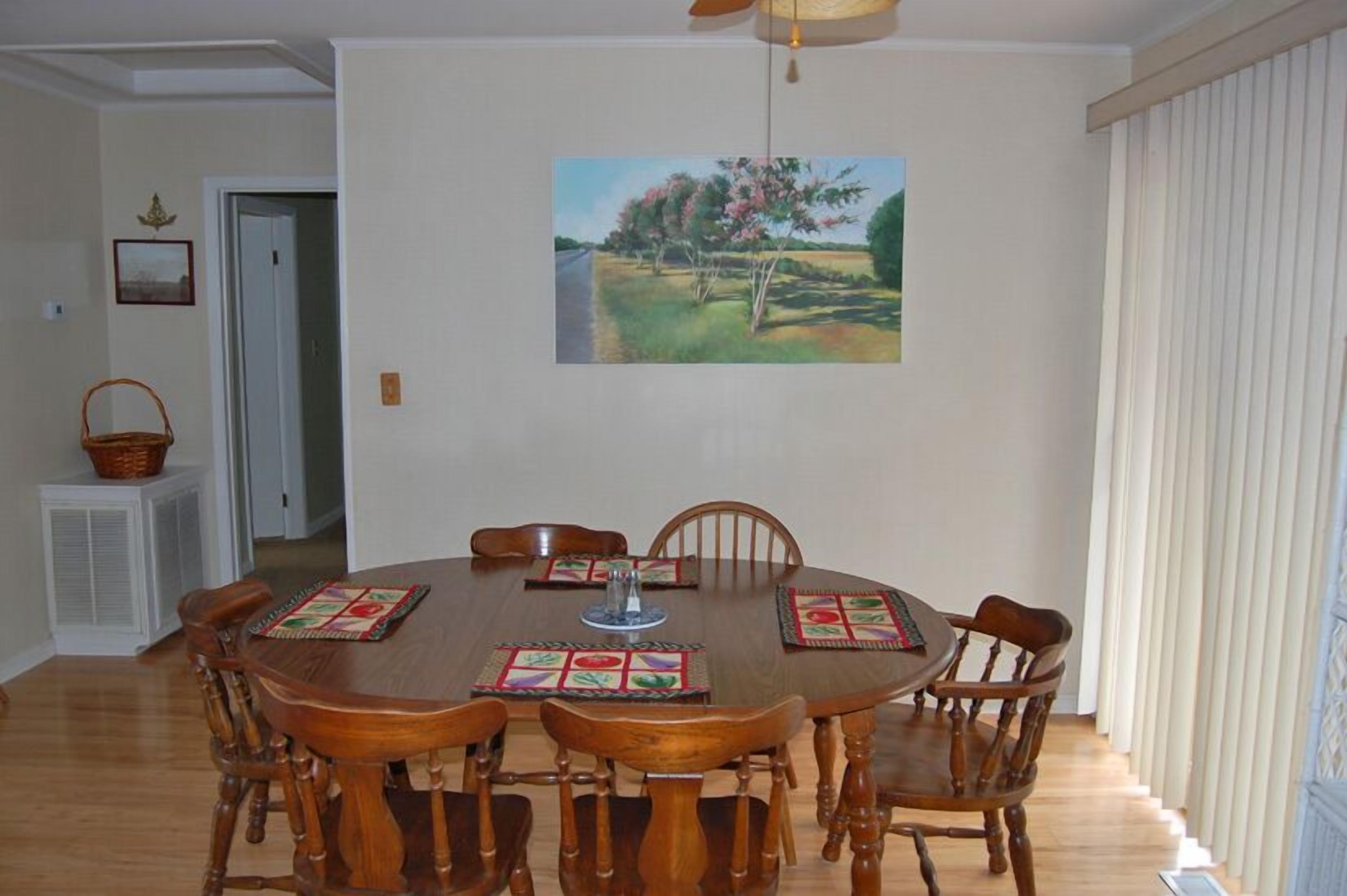Uma sala de jantar com uma mesa de madeira redonda, seis cadeiras e um quadro de uma paisagem na parede.