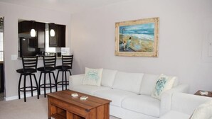 Un salon avec un canapé blanc, une table basse en bois et des tabourets de bar noirs. Un tableau représentant une scène de plage est accroché au mur.