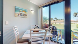 A dining area with a table and chairs, a painting of a beach scene, and a view of a lawn and palm trees through a large window.