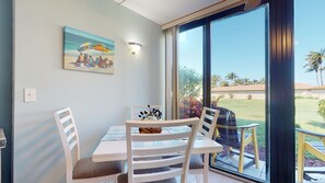 A dining area with a table and chairs, a painting of a beach scene, and a view of a lawn and palm trees through a large window.