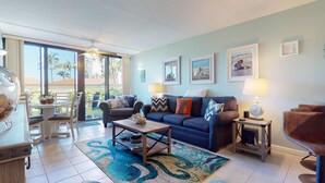A spacious living room with a large sofa, a coffee table, and a dining area with chairs. There are framed pictures on the walls and a view of palm trees outside.