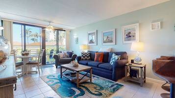 A spacious living room with a large sofa, a coffee table, and a dining area with chairs. There are framed pictures on the walls and a view of palm trees outside.