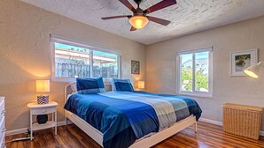 Une chambre à coucher comprenant un lit, deux tables de chevet, un ventilateur de plafond et deux fenêtres.